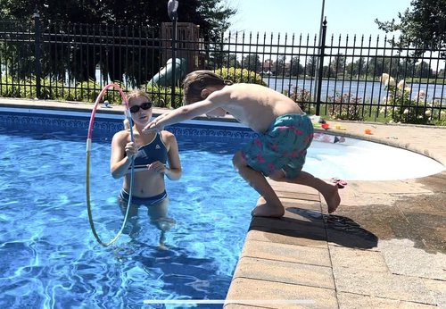 Un jeune garçon au maillot de bain bleu et motifs roses saute dans un cerceau lors d'un cours de natation à domicile avec sa monitrice