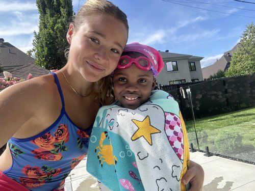Une monitrice au maillot bleu et fleurs pose avec son élève qui porte des lunettes et un masque pour cheveux roses au bord de la piscine
